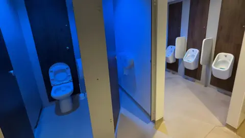 Paul O'Gorman/BBC Modern toilet cubicle with blue LED lighting close to refurbished urinals.