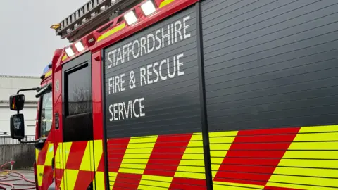 The side of a fire engine. Staffordshire Fire and Rescue Service is written on the side of the vehicle. In the distance, some pipes can be seen trailing on the ground.