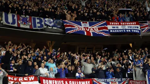 EPA Rangers fans at the game against Nice, many dressed in replica kits. Many of the fans sing and sing, while around their section there are banners indicating which area they are from. 