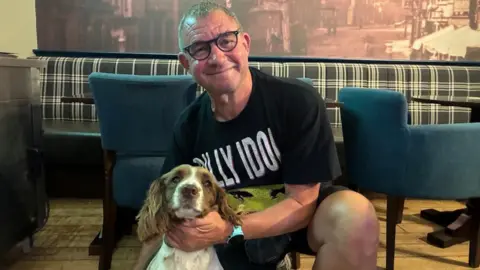 An older man with short grey hair and thick black-rimmed glasses kneels down with his arm around tan and white spaniel dog.