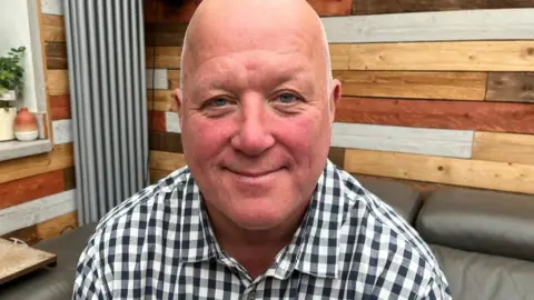 Jason is sat in his conservatory at home, in a dark leather chair. He is wearing a black and white checked shirt. In the background is multi-tone wood panelling, and a grey radiator on the wall, as well as plants on a shelf.