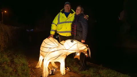 Photograph and Steve and Angela Hale stood behind a lantern otter which stands as tall as their thigh. The otter is illuminating the area around them and holds a fish in its mouth. Steve wears a bright yellow high vis jacket, has glasses and a cap on, while Angela wears a navy guilted jacket, tartan scarf and has brown shoulder left hair. 