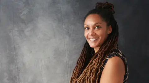 A woman with long braided afro hair is smiling as she is standing next to a grey backdrop.