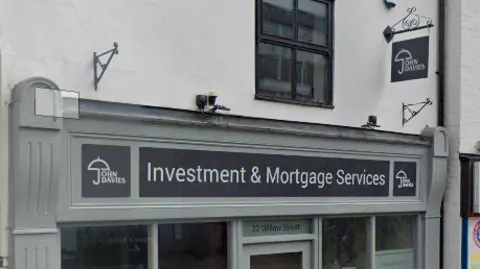 Google The front of a shop which is painted green grey and has a black and white logo. The logo says "John Davies investment and mortgage services". The wall of the shop, above the sign, is white. There is a black wrought iron hanging sign with a white "John Davies" logo.