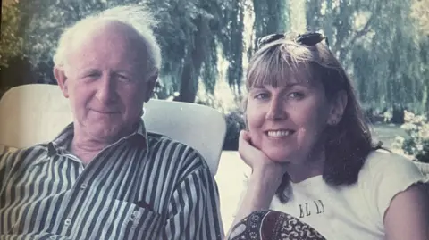 Jane Arnold An old photograph with faded colours of Jane Arnold smiling with her father Barry on a sunny day in a garden. Jane has blonde hair and has sunglasses on her head. Barry has white hair and a striped shirt.