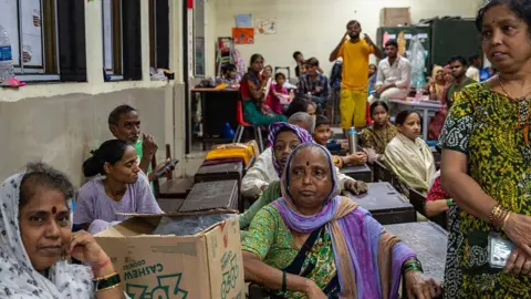 Hindustan Times via Getty Images women sitting on a bench in a class that has been transformed into a temporary shelter. The cardboard box lies next to one of the women who wears green sarai and is in the center of the image. 