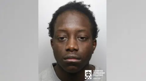 A mugshot of a young man with short black hair and wearing a grey jumper.