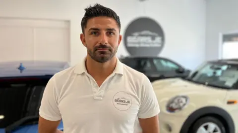 Tiago is standing in a car showroom, wearing a white polo shirt with the logo 'SEARS-JE Since 1993' on the left chest. Behind him are several cars, including a blue one on the left and a white one on the right. A circular sign on the wall reads 'GEARHOUSE' with a car graphic above it.