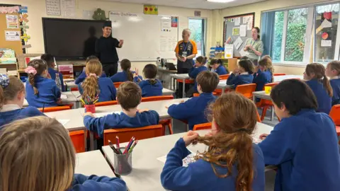 Chew Stoke Church School Lando is standing in front of a classroom full of children. He is giving a talk about his career. The children are facing him and wearing blue uniform and sat on red chairs. Two school teachers are standing next to him. 