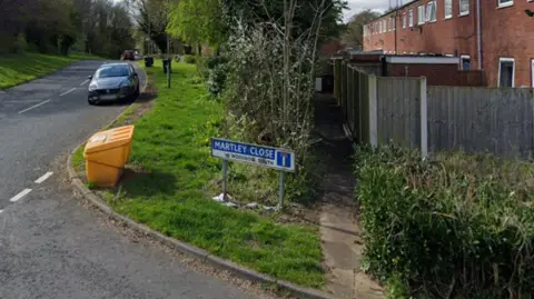 A blue and white road sign reading Martley Close on grass on the side of a road junction. A car can be seen in the distance and hedges, a fence and a building behind.