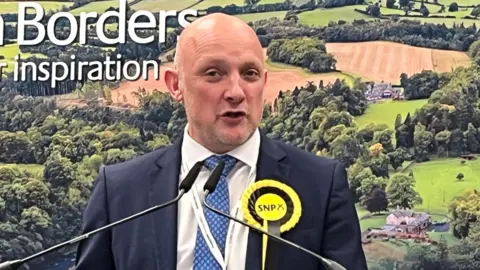 Callum Kerr - a bald man in a suit with an SNP rosette - stands in front of photograph of the countryside, speaking into two microphones. 