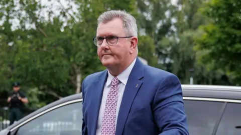 Jeffrey Donaldson is walking away from a black car.
He is a man with short grey hair, wearing a dark blue suit, white shirt and pink tie.