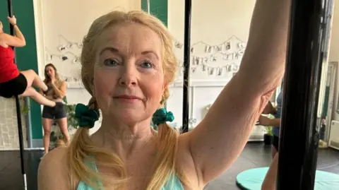 Karen is holding onto a vertical pole inside a fitness studio designed for pole exercise. The studio has light-colored walls with a green accent panel and a display of photos or notes arranged on strings in the background. Other individuals are visible practicing on poles, and a round exercise mat is placed on the floor.