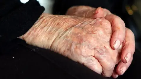 PA Media An elderly lady's hands clasped together with chipped nail varnish and black clothing
