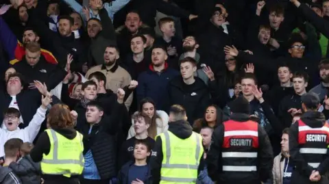 A crowd of Southend United FC fans in a stadium, men women and children. Facing them with their back to the viewer are two stewards in yellow high-vis and two men with security on their backs. 