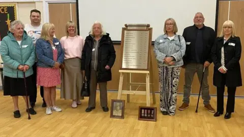 A group of relatives are stood in a line to the left and right side of the newly unveiled war memorial. The memorial lists fifteen men who lost their lives in World War II. In front of the memorial there are two frames containing a photograph of one of the men and his medals.
