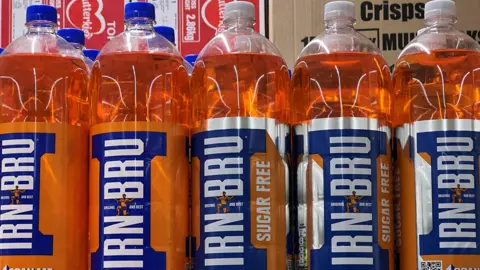 Two litre bottles of Irn-Bru on a supermarket shelf. The liquid is orange in colour and the labels on the bottles are blue and orange with IRN-BRU in white letters.