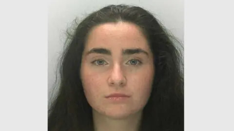 A police mugshot of Liberty Mitchell. She has thick eyebrows, long dark hair and grey-blue eyes, and is looking right at the camera with a blank expression.