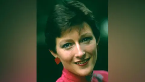 Sally is wearing a pink top with white detail and a high collar around her neck. She has large red diamond-shaped earring and short dark hair, 