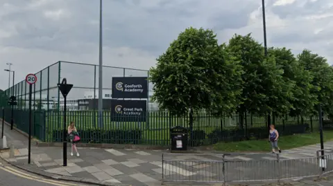 A green fence surrounds a playing field. A black and gold sign says: "Gosforth Academy". 