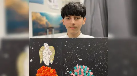 A teenage boy with mid-length black hair holds up a canvas that appears to show an angel in a night sky