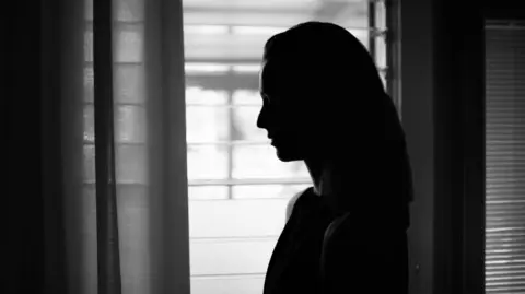 A woman stands silhouetted beside a window, her features obscured by shadow and fading light.