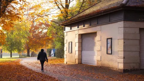 Mark Pemberton A person wearing headphones walks along a leaf-strewn pathway past a park building towards a row of trees showing autumnal colours