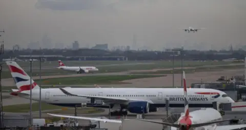 Reuters Planes arriving and departing from the runway at London Heathrow. One plane is coming into land, whilst others traverse the runways on the ground, where a BA plane parks at a gate whilst another smaller Swiss Airline plane also taxis behind it. The London skyline is clearly visible in the distance on a grey cloudy day, with the Shard being especially prominent.