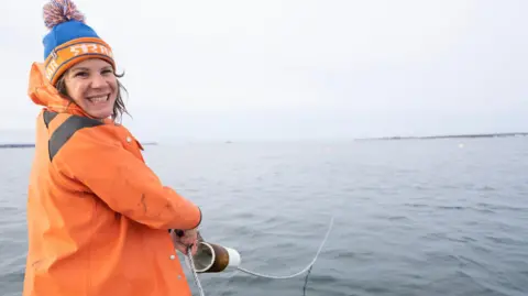 Elizabeth Ellenwood Kelp farmer Suzie Flores wearing an orange waterproof jacket and orange and blue bobble hat, with the sea behind her