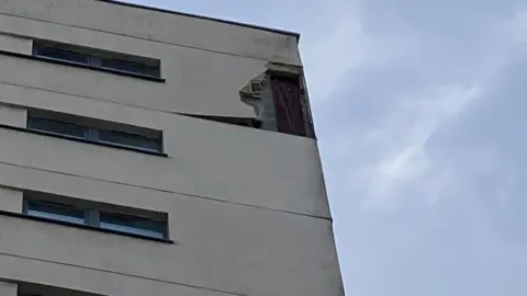 A high-rise building seen with window. There is a large chunk missing from the corner of the building with the panelling seen inside it.