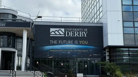 University of Derby, Kedleston Road campus. It is a large white building with a large banner in front with the University of Derby logo and the words "The future is you" underneath
