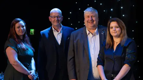 Wiltshire Council Four people dressed smartly - two men and two women - look at the camera standing in front of a dark but lit backdrop