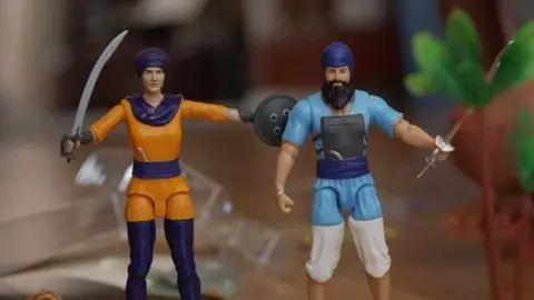 Two toys stand on a table - a female character in orange and purple clothing with a sword and shield and a male character in blue clothing with a sword. Both wear turbans