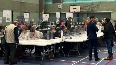 Counters go through ballots at a previous Cherwell District Council election count. They sit at white trellis tables in a sports hall, behind plastic screens. Candidates and members of political parties watch the count. 