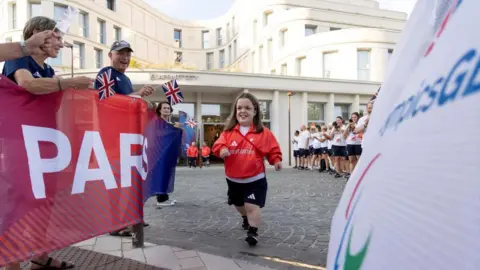 ParalympicsGB Charlotte McGuinness wearing a red Great Britain jacket and dark shorts is moving forward along a paved courtyard area, with people on both sides cheering and holding small Union flags. On the left, spectators stand behind a large red banner. On the right, part of a white banner with the Paralympic symbol is visible in the foreground.
