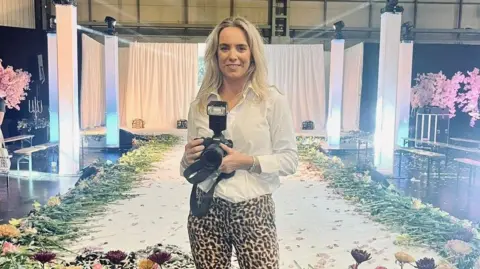 Gina Gray A lady standing in front of a catwalk that has flowers laid along the edge. She is wearing leopard print trousers with a white shirt and is holding a professional camera. The lady, who has shoulder length blonde hair is smiling. 