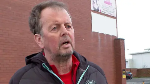 A man stands at an angle, talking to someone unseen while standing in front of a brick wall. He has dark hair and is wearing a padded black jacket, red t-shirt and red tracksuit top he has stubble down his face.