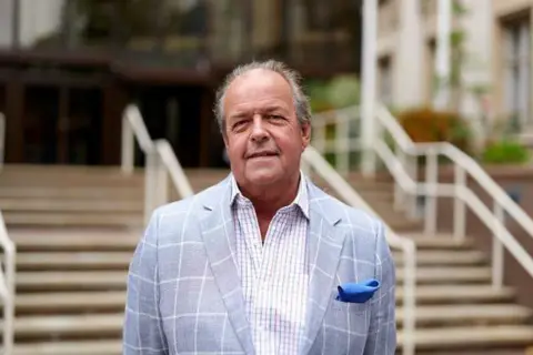 Essex County Council John Spence has brushed back hair, and wears a light blue checked jacket with a bright blue handkerchief in the top pocket. He is wearing a shirt with a smaller white and blue check underneath the jacket and is standing in front of steps.
