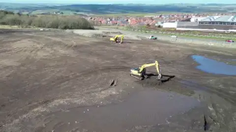 Project Genesis Two yeallow diggers at work on the vast muddy site of the new hospital. They are surrounded by two large puddles. The land is near a road. Some housing can be seen across the path.