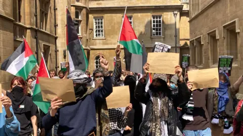 Mousumi Bakshi/BBC Demonstrators in Cambridge can be seen waving Palestinian flags and signs. They wave their hands, flags and signs in the air as they march.