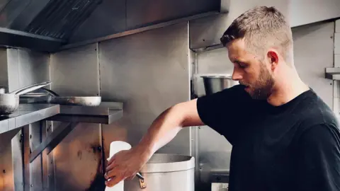 The Social Trattoria Justin Ashwin pours a bottle into a pan on his restaurant kitchen cooker. He wears a dark shirt.