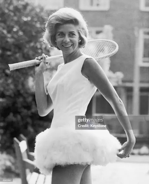 Getty Pericoli holds a racquet and wears a white dress trimmed with feathers