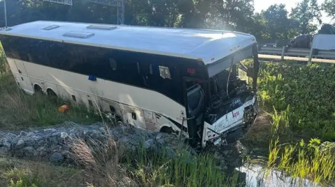 A white and black bus sits on greenery on the side of a road after a crash.