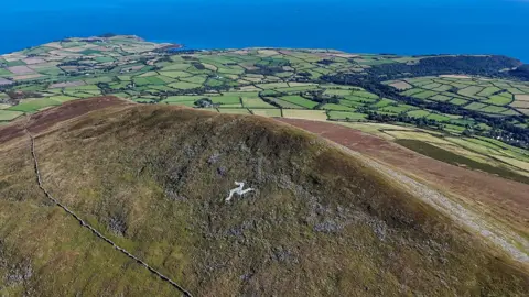 Manx Scenes A tall ridge of a grassy hillside stretches from the right to the middle centre of the view, with a mass of green fields below and the sea beyond. On the ridge of North Barrule a white Three Legs of Man symbol is clear, the shot taken on a sunny day.
