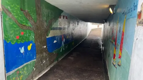AlexPope/BBC An underpass, showing old worn paint, and a path, that is dark. The paintings on the wall include a tree, boats and colourful images. 