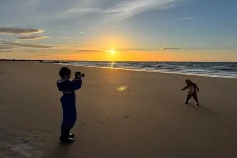 Zoe Mann Two young boys on a beach at sunset, one is holding a camera, the other is making a dance shape, the sun is glowing in the distance over the sea.