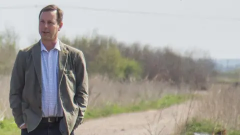 Holly Wilkinson Matt Jackson, standing in a nature reserve, wearing jeans, with a belt, a checked shirt, and jacket, looking slightly away to the left.
