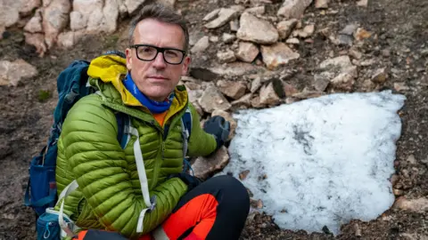 Snow expert Iain Cameron, wearing glasses and a great rain jacket, poses next to the Sphinx snow patch, which is about one metre wide and is melting into the ground below.