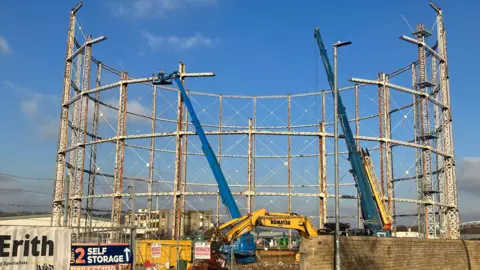 A gas holder in the process of being pulled down, with a blue crane and a blue cherry picker being used to demolish the structure. Blue sky with little cloud cover can be seen in the background. 