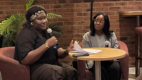 A woman holding a microphone and a pile of papers sits on a chair at a small desk. Another woman holding a microphone sits in the chair next to her. A brick wall and a plant can be seen behind her.